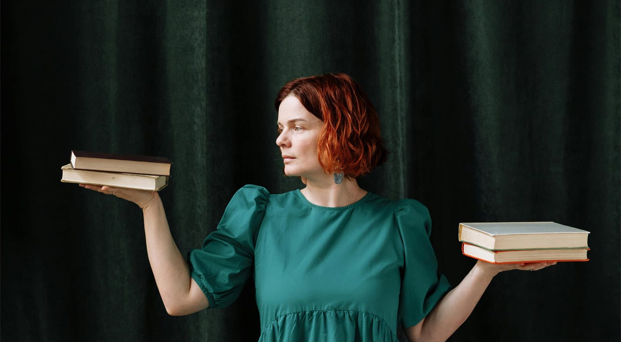 Woman standing against a green curtain, balancing two books in the palm of each hand - looking like she's weighing them up